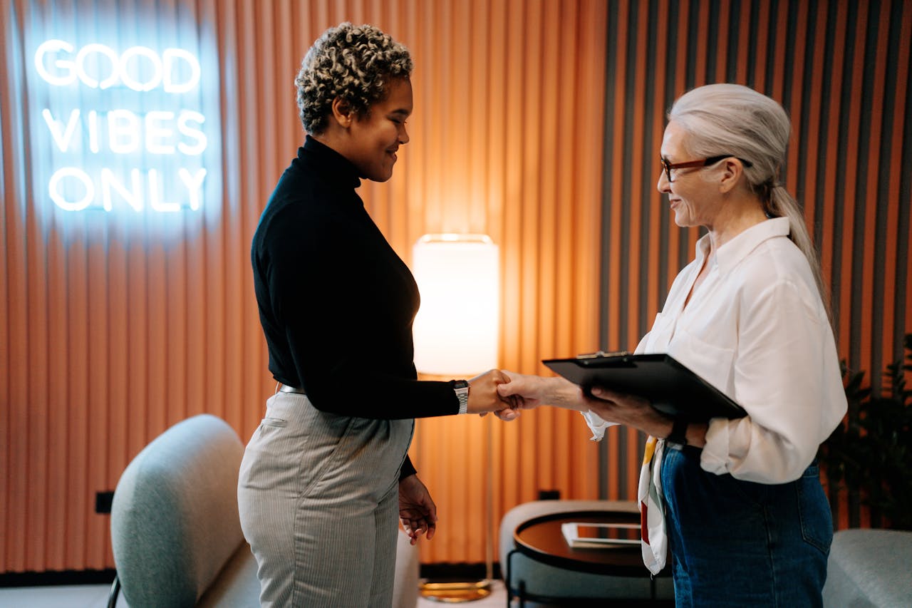 impact-img Two diverse businesswomen in a stylish office shaking hands under a Good Vibes Only sign.